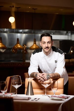 Tattooed chef in white coat poses at elegantly set restaurant table with wine glasses and copper pendant lights.