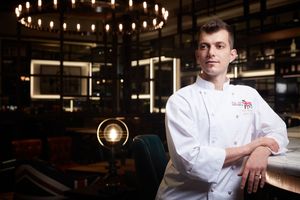 Professional chef in white uniform poses confidently in an upscale restaurant with elegant chandelier lighting.