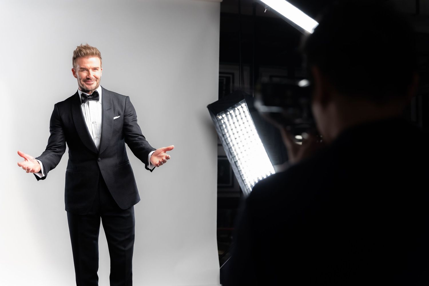 Man in black tuxedo poses for professional photo shoot with studio lighting and photographer in background.