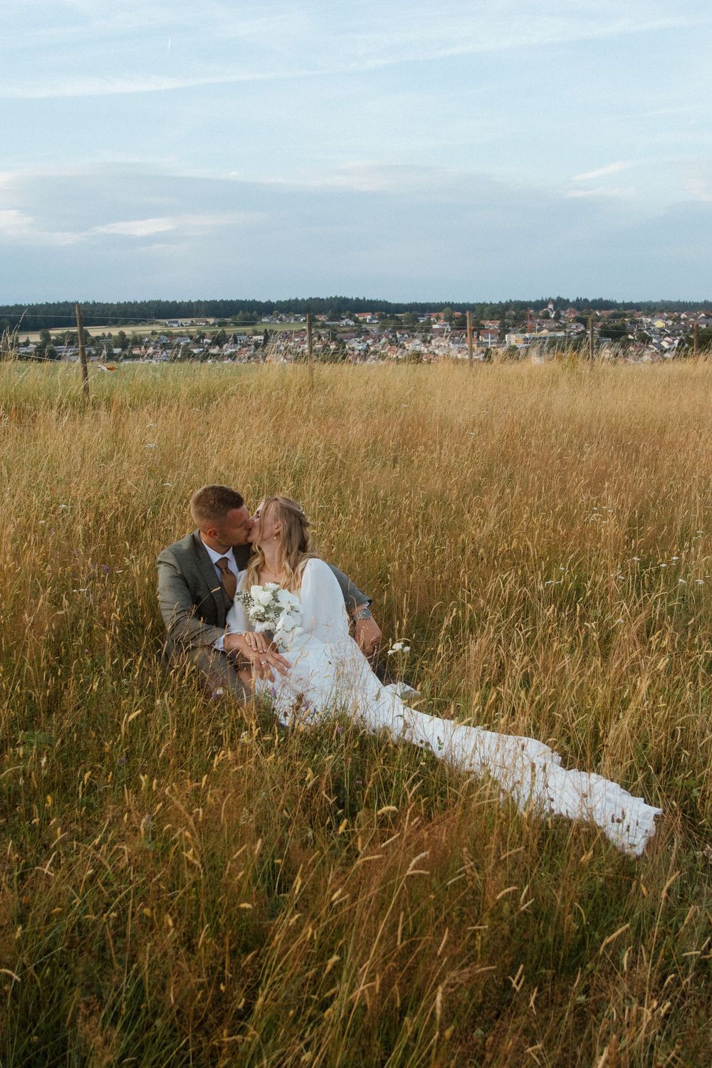 A couple shares a romantic moment in a golden grass field with a distant town and mountains in the background.