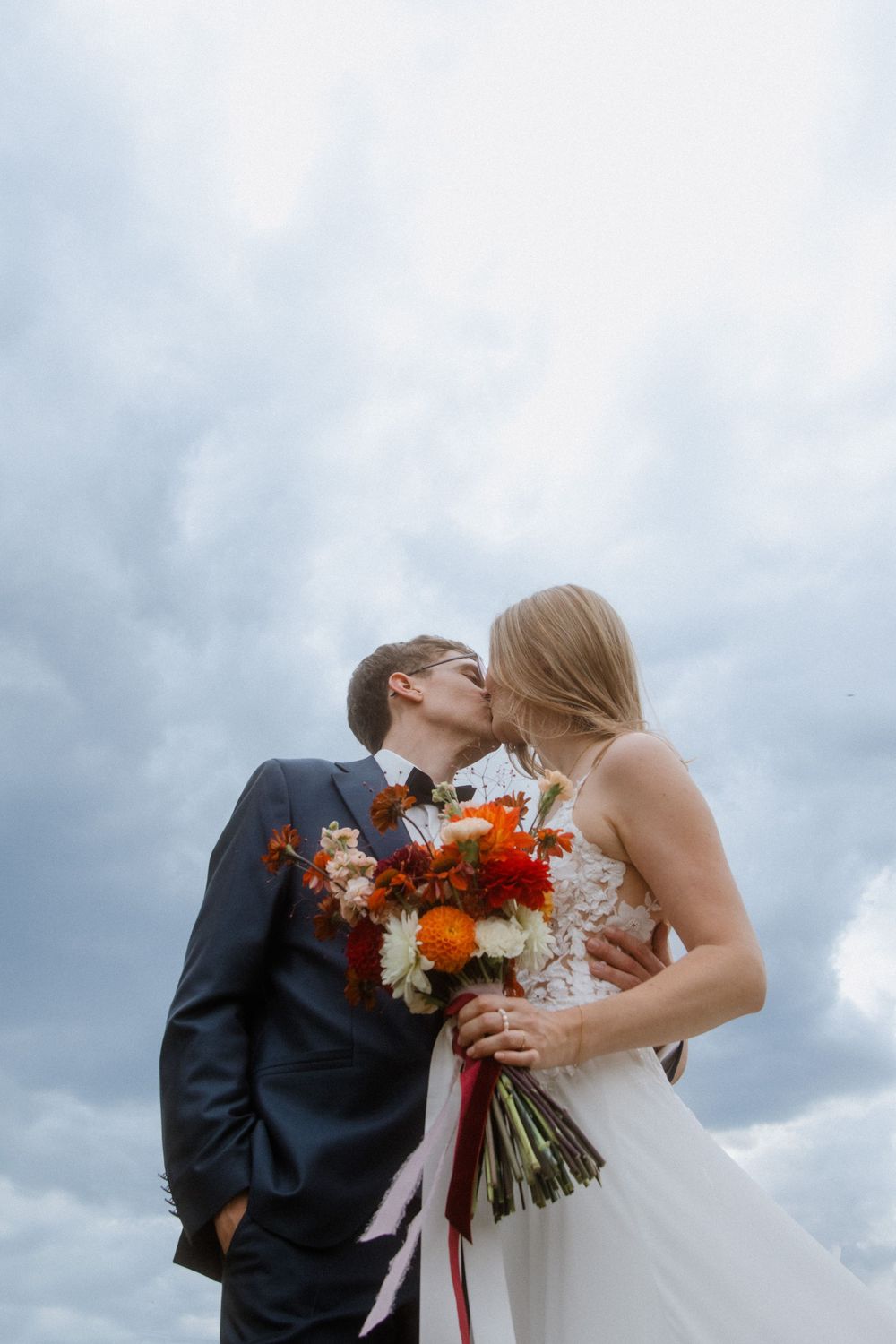 A romantic wedding couple shares a tender moment against cloudy skies with a vibrant autumn-colored bouquet.