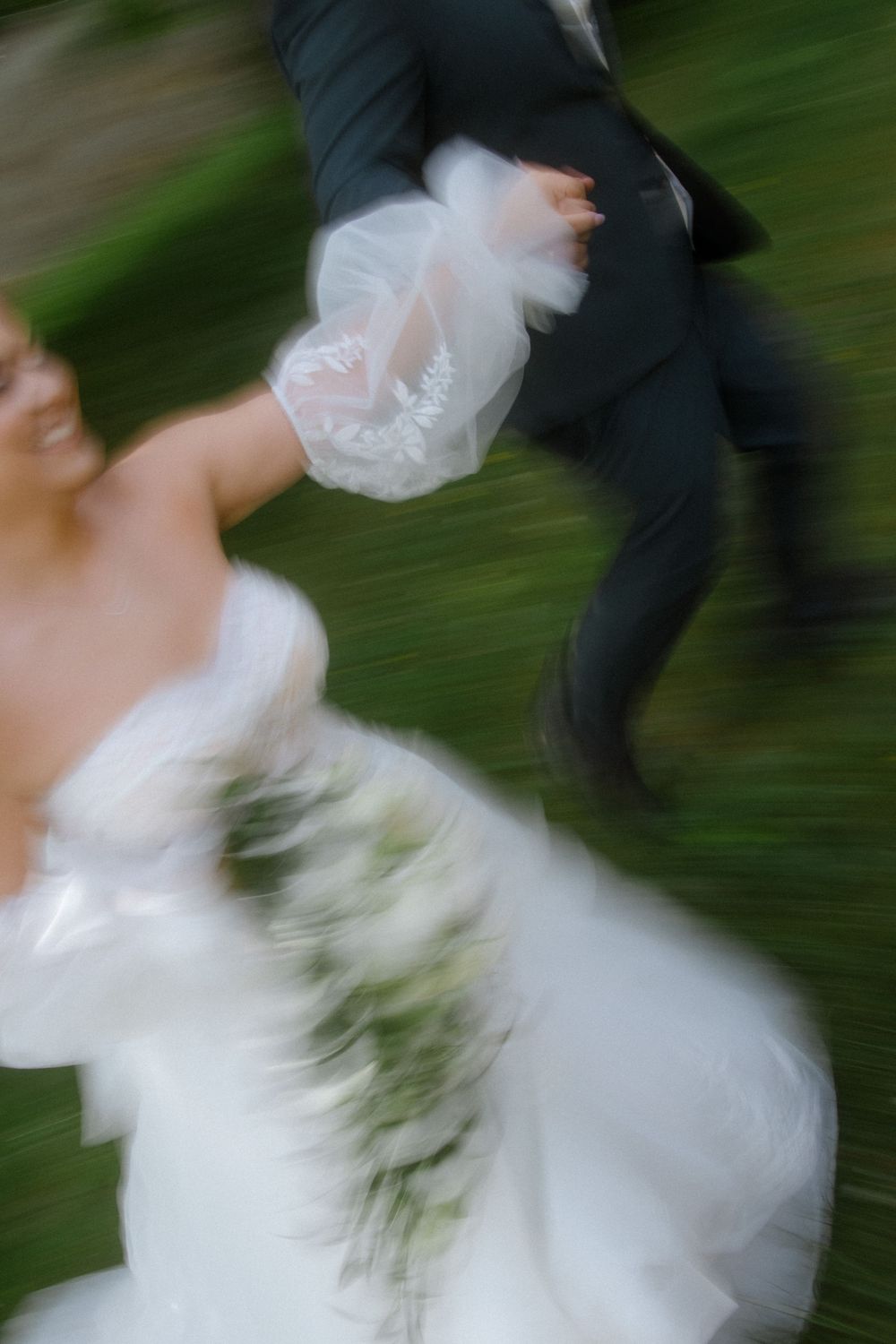 Motion-blurred artistic wedding photo shows couple dancing, with bride's white dress swirling against green background.