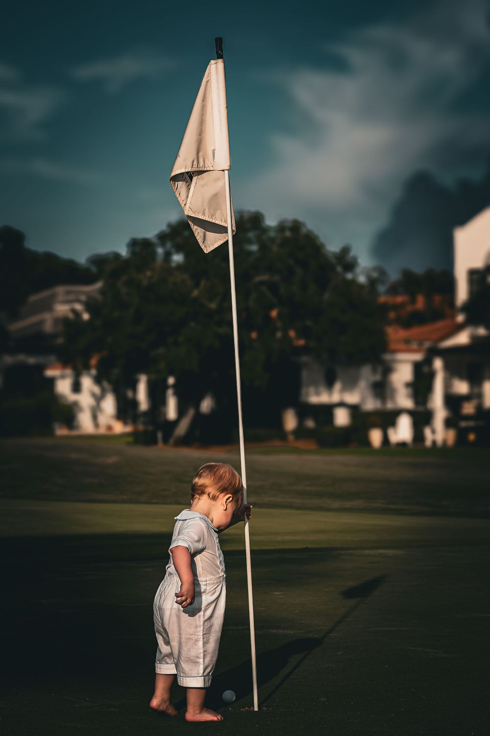 A small figure stands beside a tall white flagpole on a golf course at dusk.
