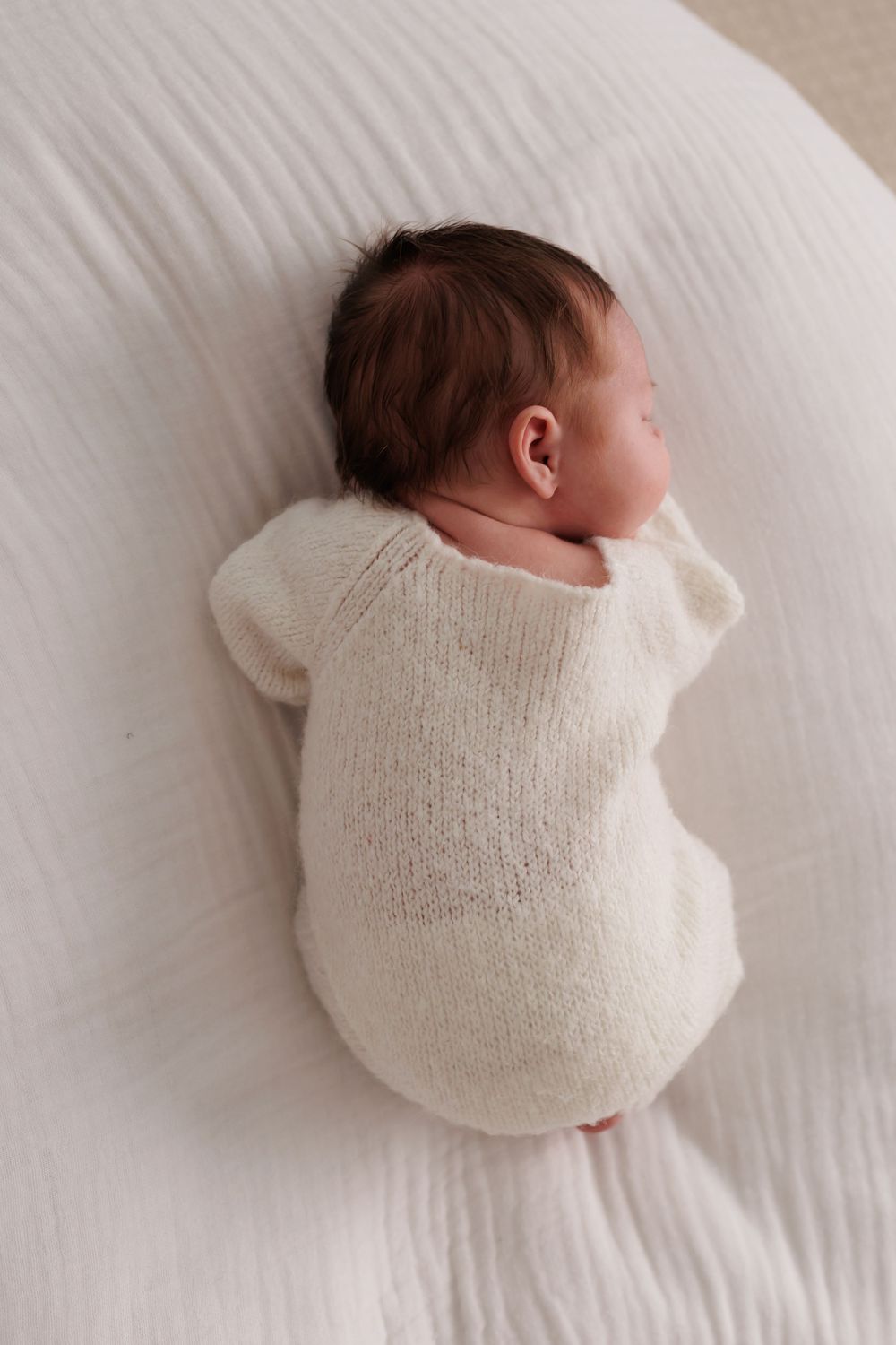 Newborn baby sleeping peacefully on white bedding, wearing a cream knit romper, photographed from above.