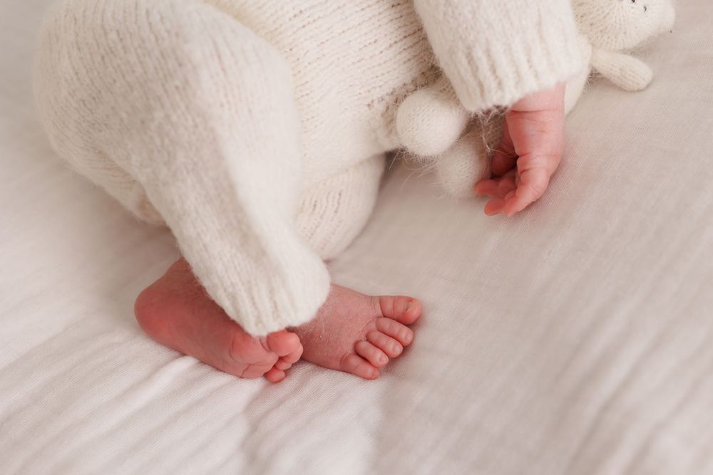 Close-up of newborn baby feet in white knit outfit lying on soft white bedding with stuffed animal.