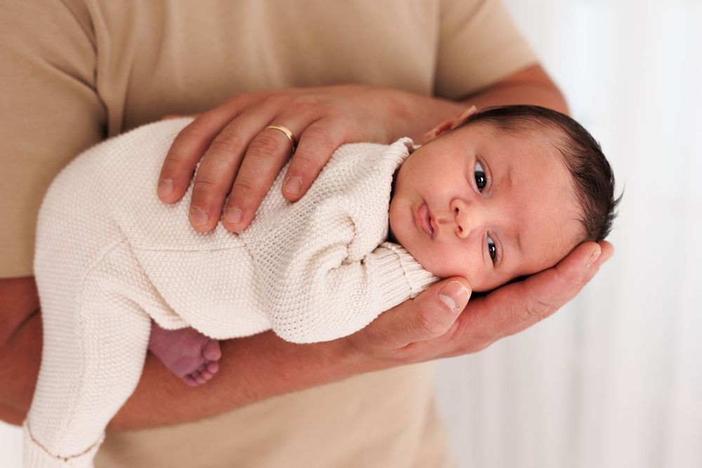 Newborn baby in white knit outfit cradled in father's hands, peaceful and alert.