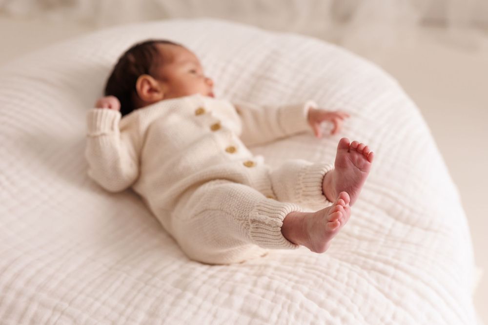 Newborn baby sleeping peacefully on white cushion wearing cream knit outfit with gold buttons.