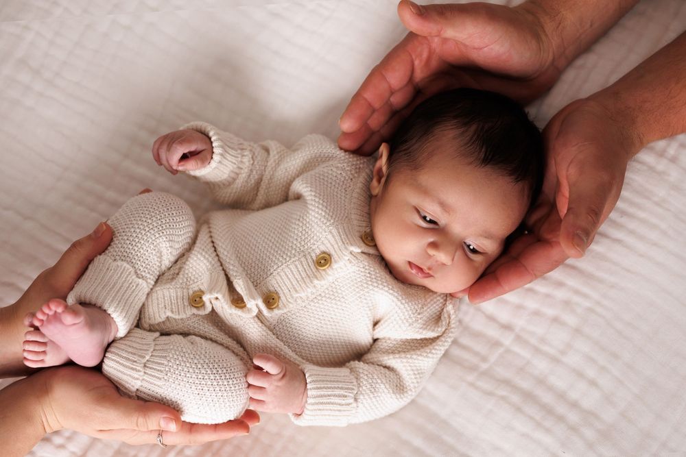 Newborn baby in cream knit outfit cradled gently by adult hands on white blanket.