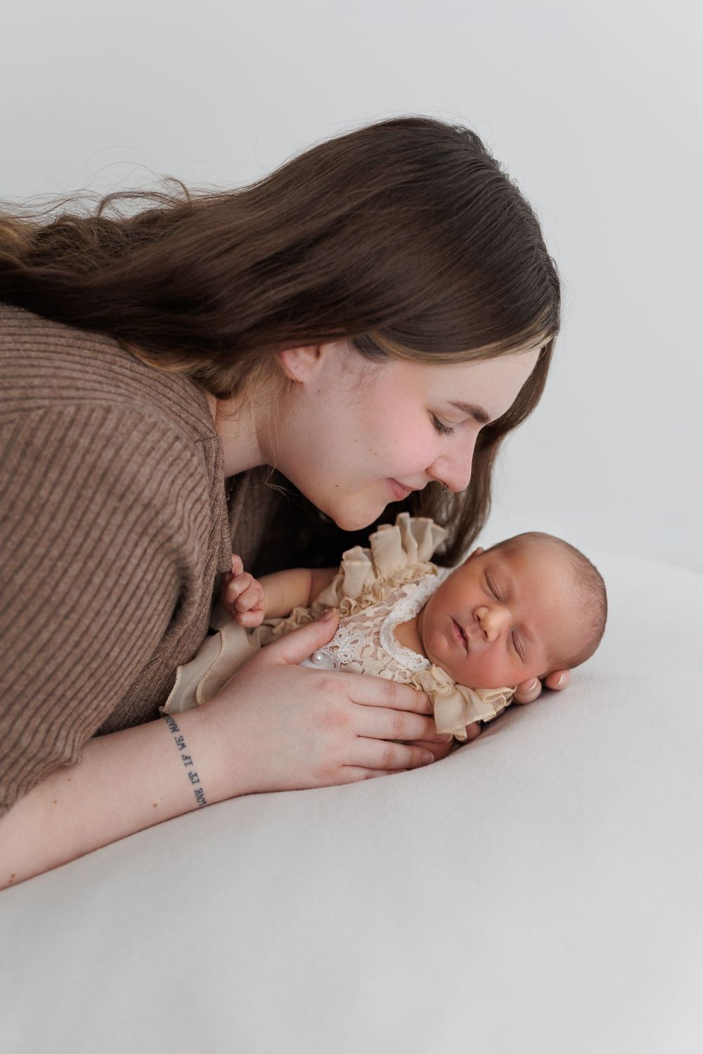 Young mother tenderly gazing at her sleeping newborn baby wrapped in cream lace, studio portrait on white background.