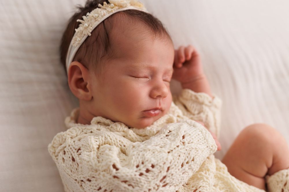 Sleeping newborn baby girl in cream knit outfit and floral headband, peaceful nursery portrait photography.