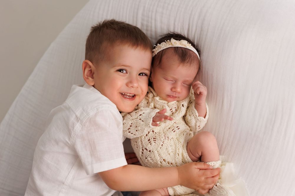Young boy in white shirt smiles holding sleeping newborn baby girl in cream knit outfit with bow headband.