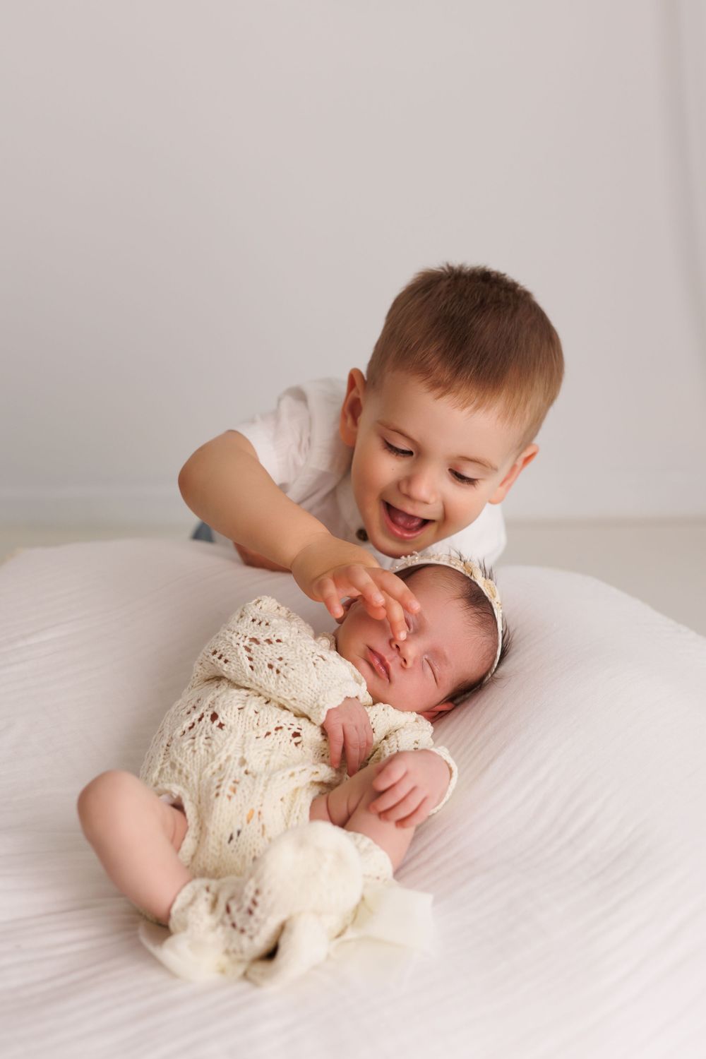 Happy toddler boy smiling while gently touching sleeping newborn sibling in cream knit outfit on white bed.