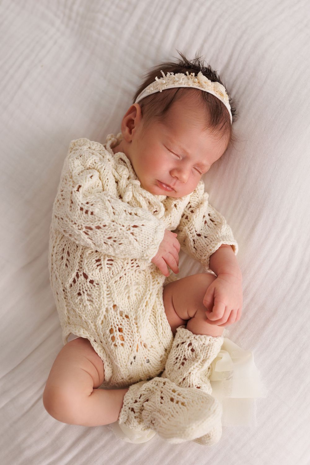 Sleeping newborn baby girl in cream knit lace outfit and floral headband lying on white blanket.