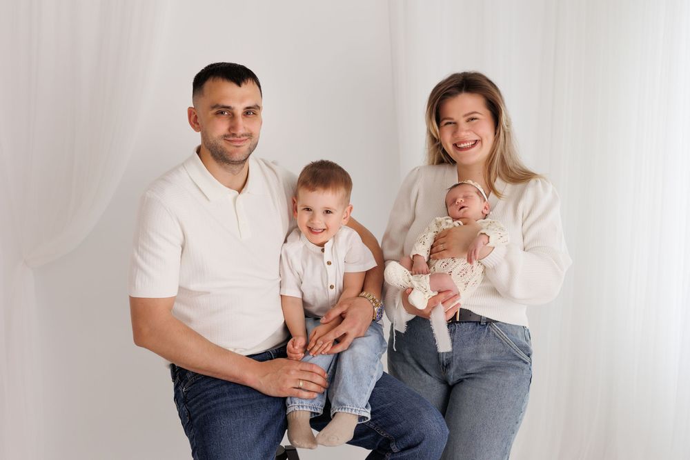 Happy family of four posing for portrait, parents with toddler son and newborn baby in bright white studio.