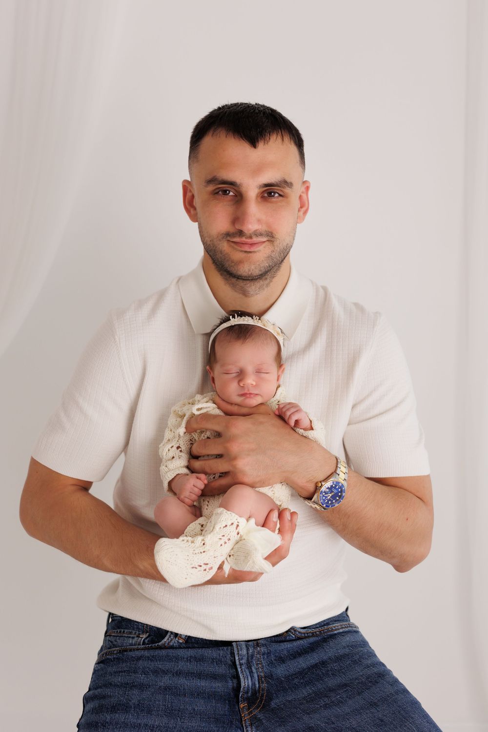 Father in white shirt holds sleeping newborn baby dressed in cream knit outfit against white background.