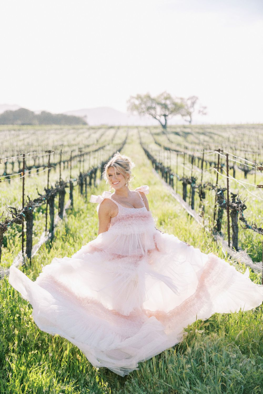Wide-angle shot of a bride in a flowing gown standing among lush vineyard rows.