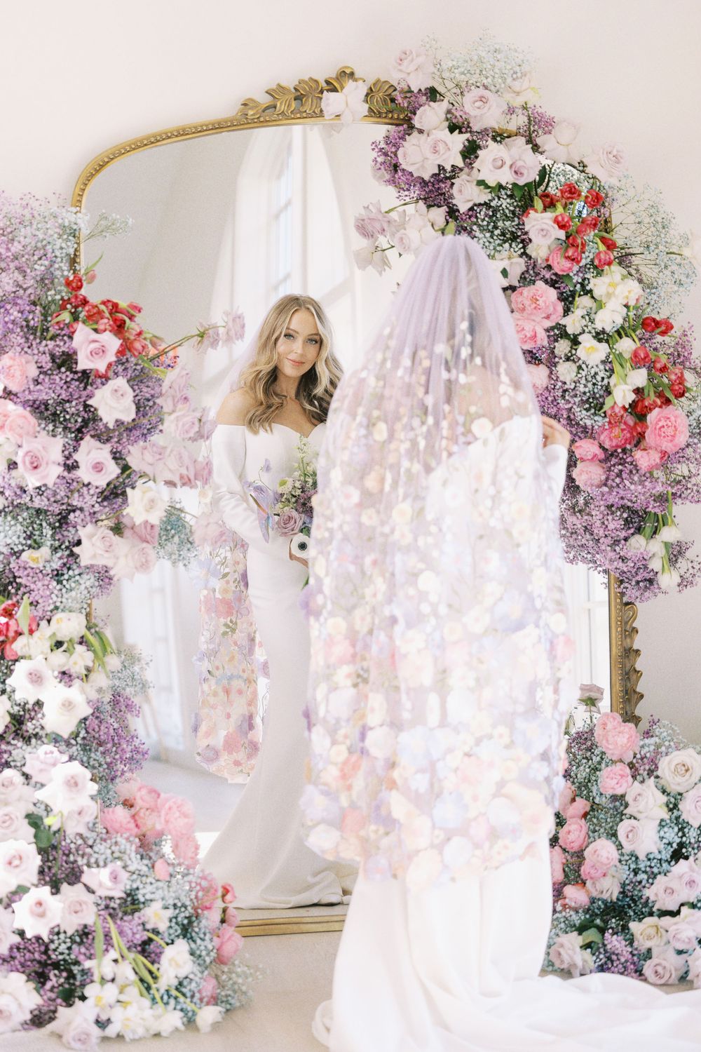 Elegant bride in gown and veil admiring herself in an ornate, gilded mirror adorned with colorful flowers.