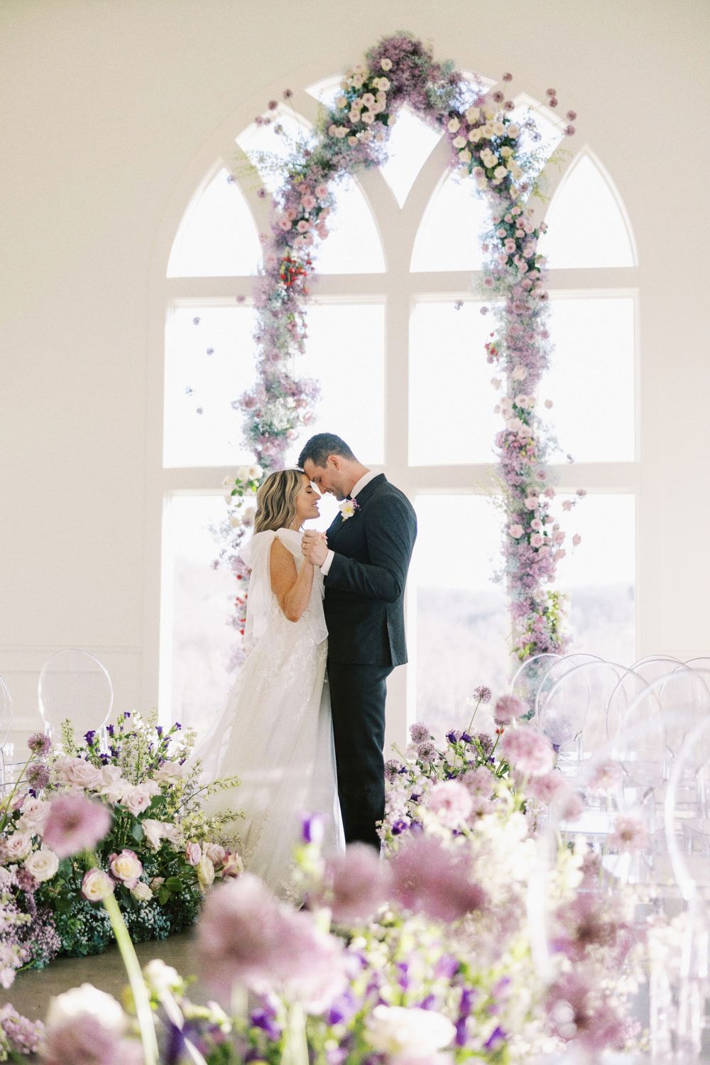 Bride and groom sharing an intimate embrace under a lush purple floral wedding arch.
