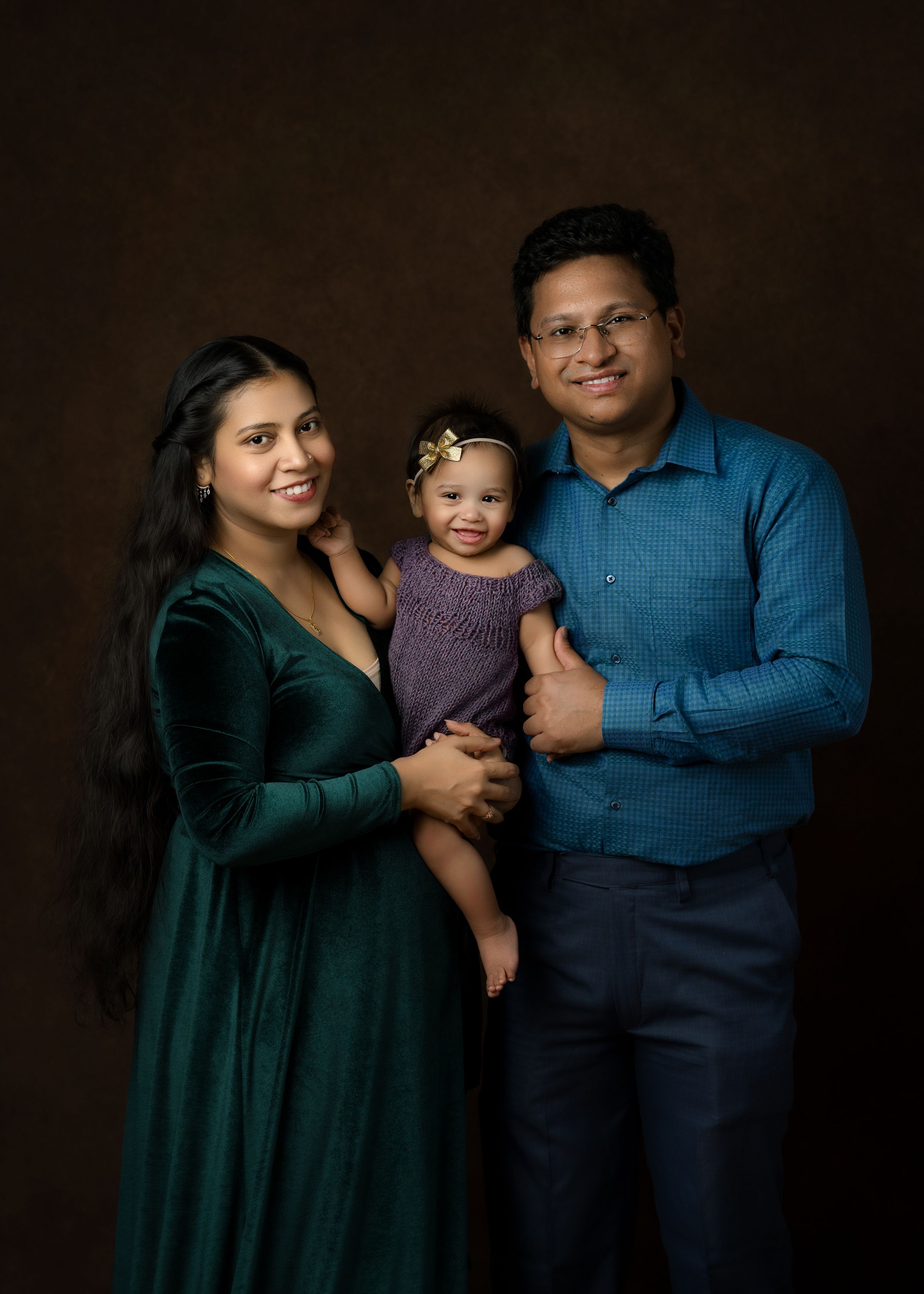 Professional studio portrait of family of three wearing coordinated emerald green and navy outfits against dark background.