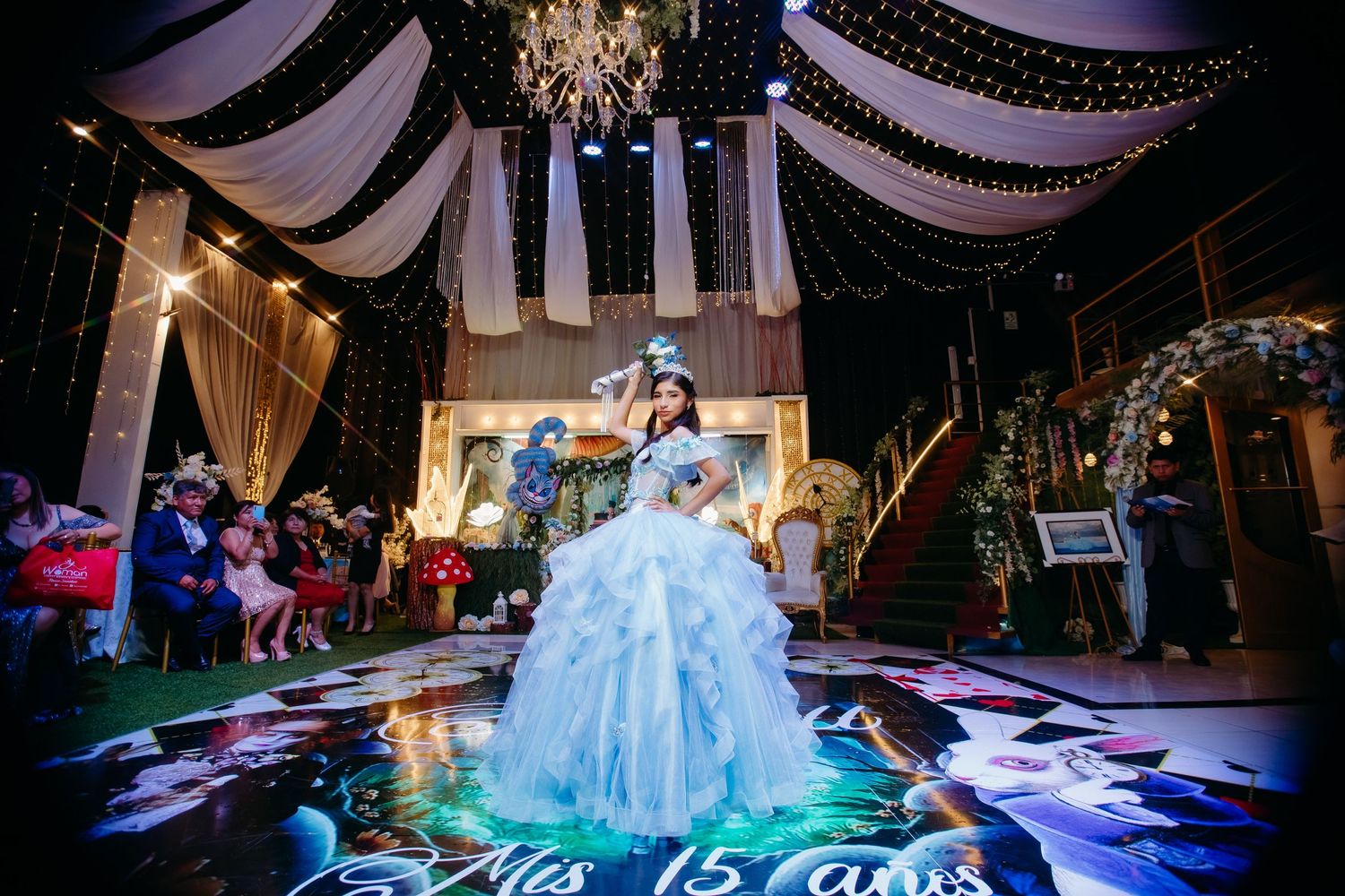 Bride in blue ball gown stands under elegant draped fabric and chandelier lighting during quinceañera celebration.