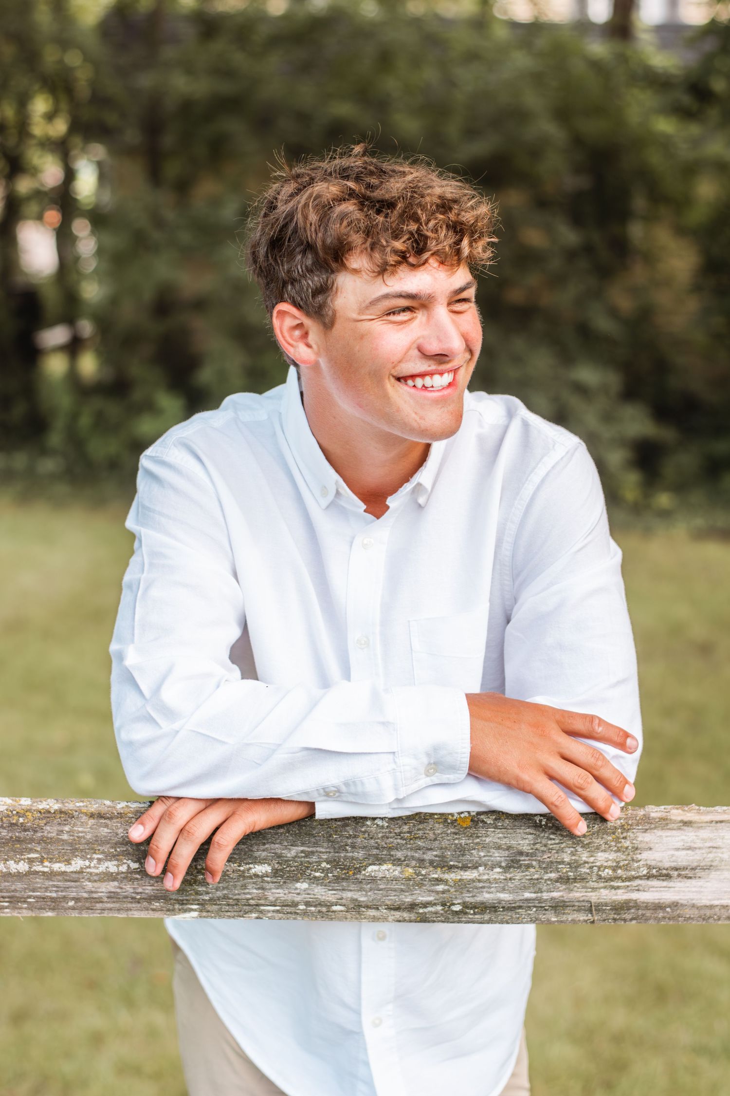 Smiling young man in a white button-down shirt leaning on a wooden fence in an outdoor senior portrait.