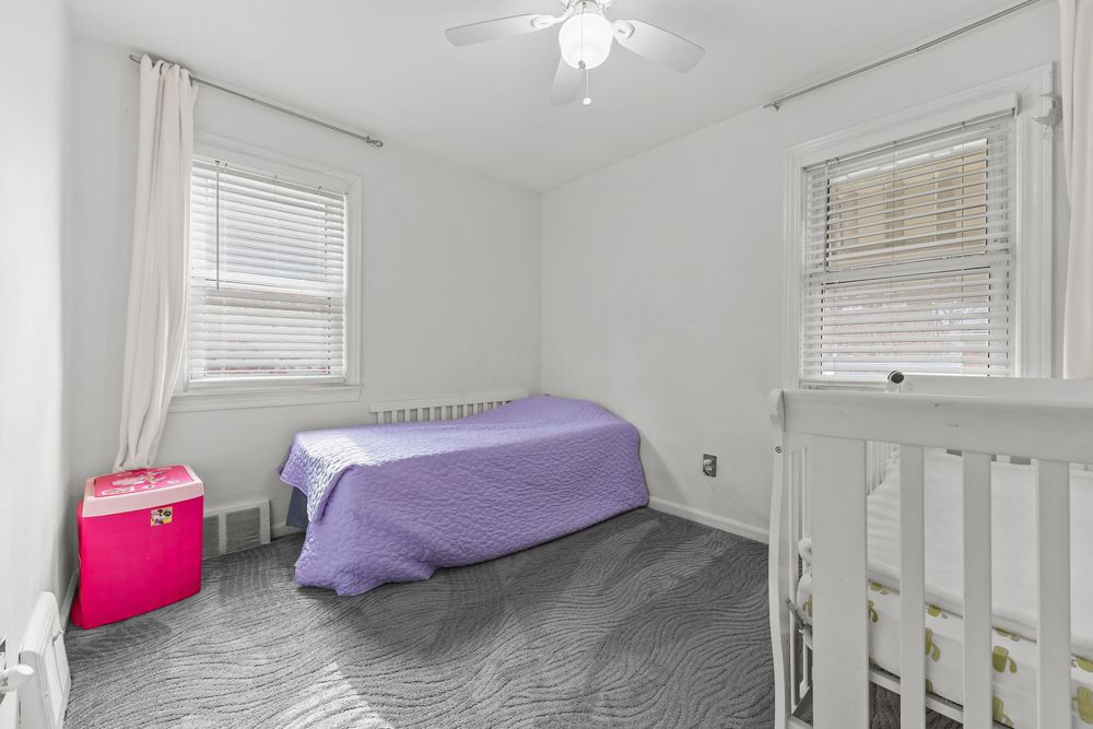 Small bedroom with gray carpet, ceiling fan, purple bedspread, and multiple windows providing natural light.
