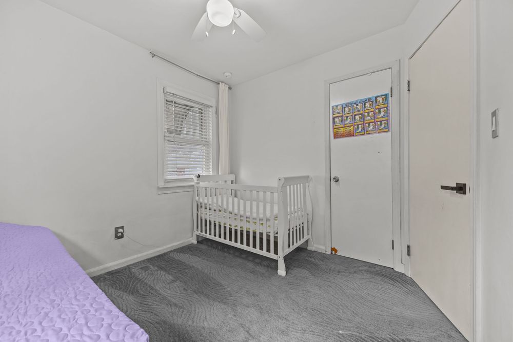 Nursery bedroom with white crib, gray carpet, ceiling fan, and closet door in a bright white room.