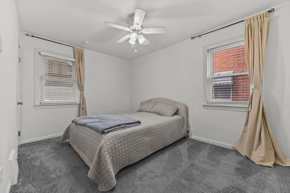 Bedroom with gray carpet, ceiling fan, tan curtains, and two windows allowing natural light to fill the space.
