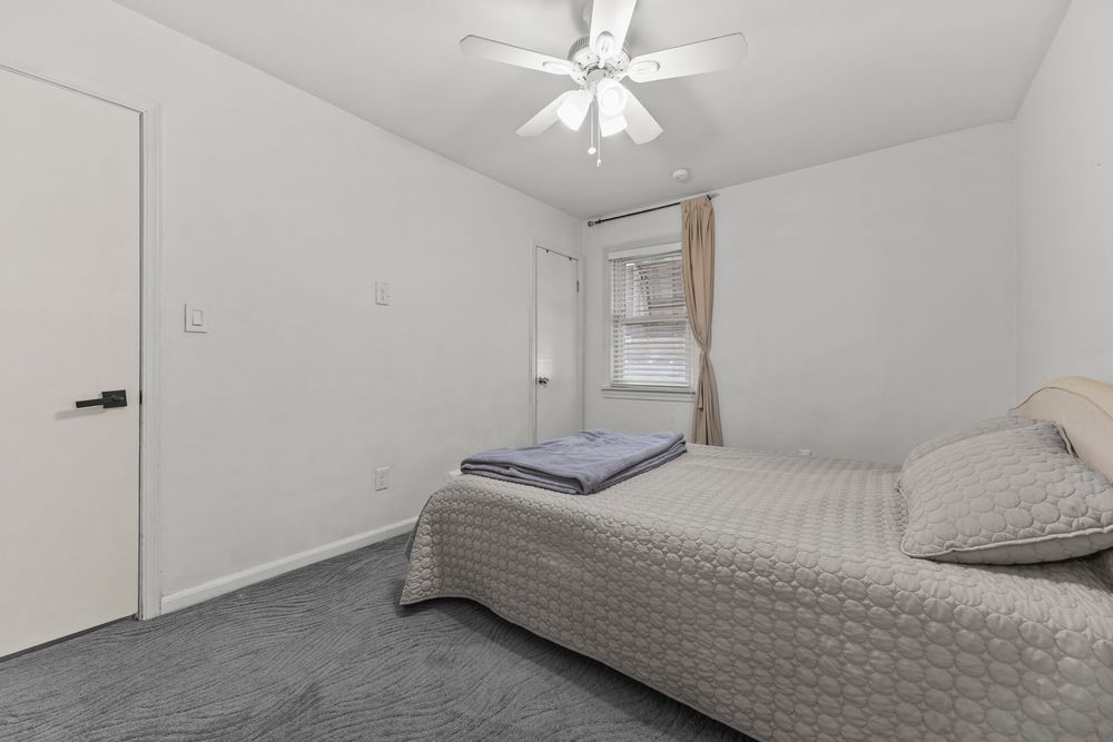 Simple bedroom with gray carpet, ceiling fan, single window with curtain, and white walls in cozy layout.