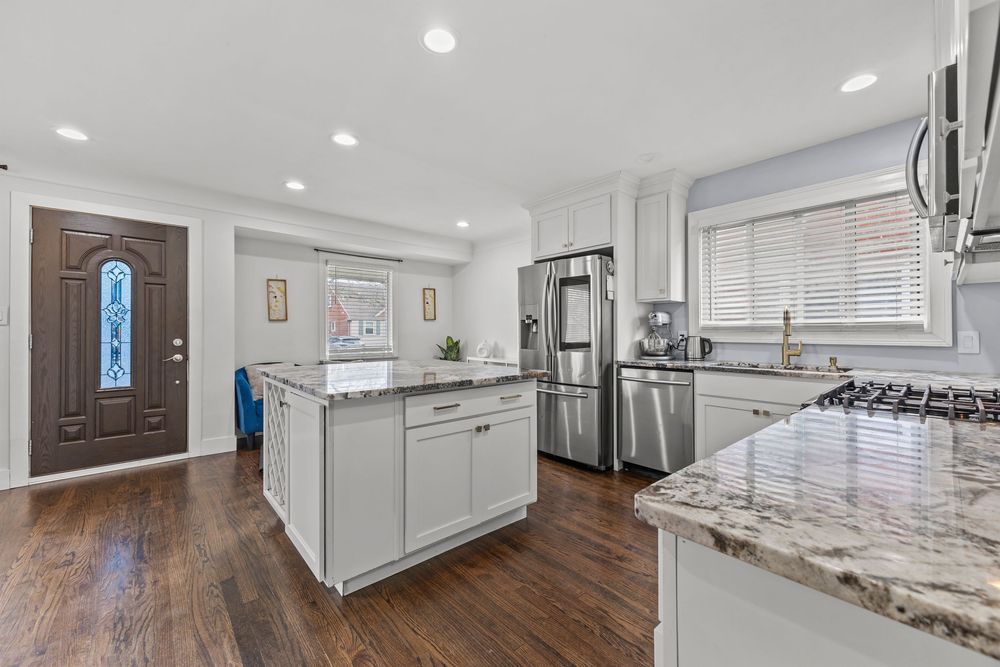Modern kitchen with white cabinets, granite island, stainless steel fridge, and dark hardwood floors.