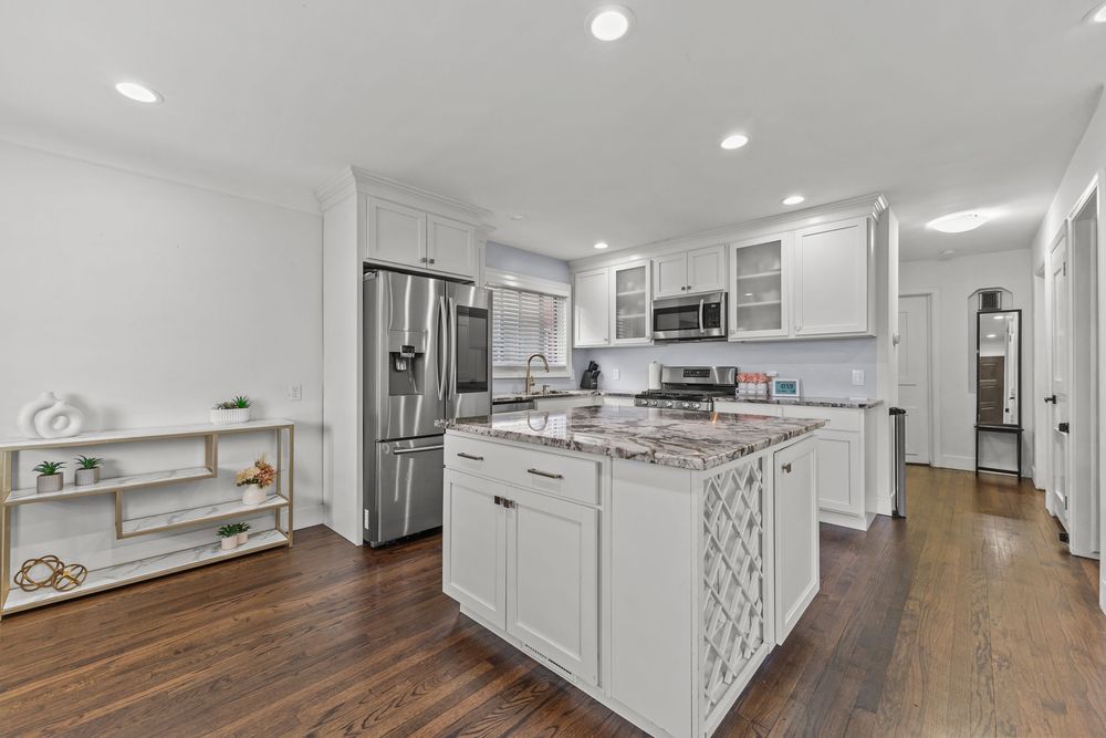 Bright white kitchen with granite island, stainless steel appliances, recessed lighting, hardwood floors.