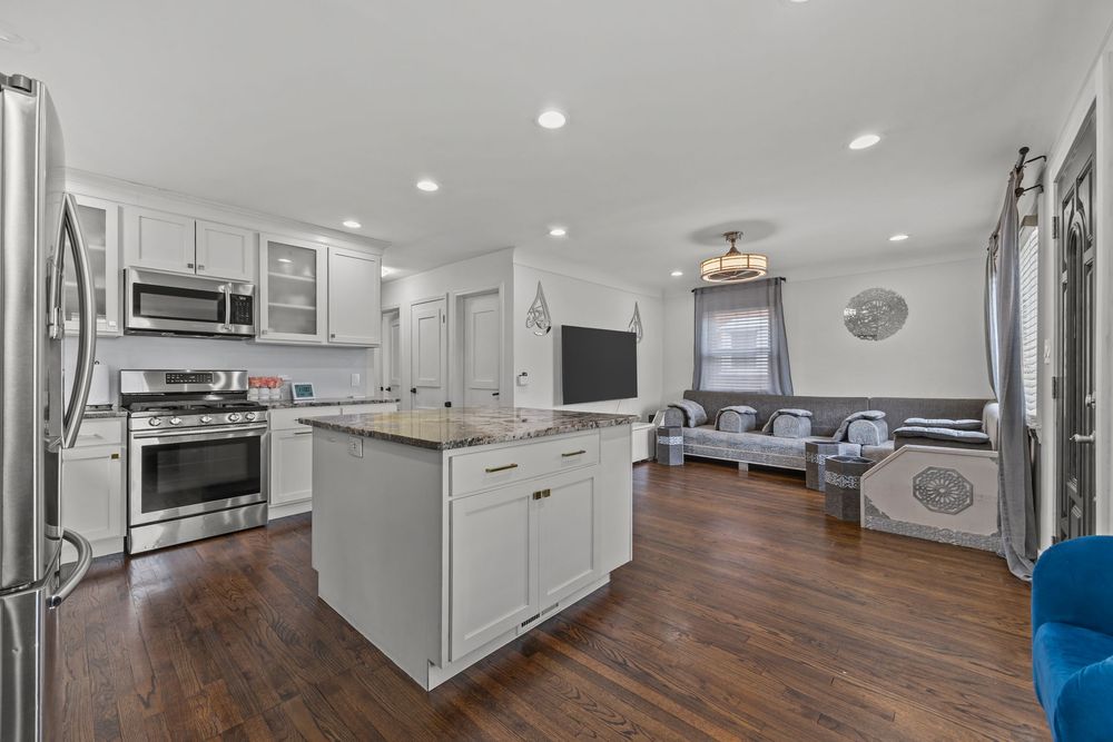 Modern kitchen with white cabinets, granite island, stainless steel appliances, and dark hardwood floors open to living room.