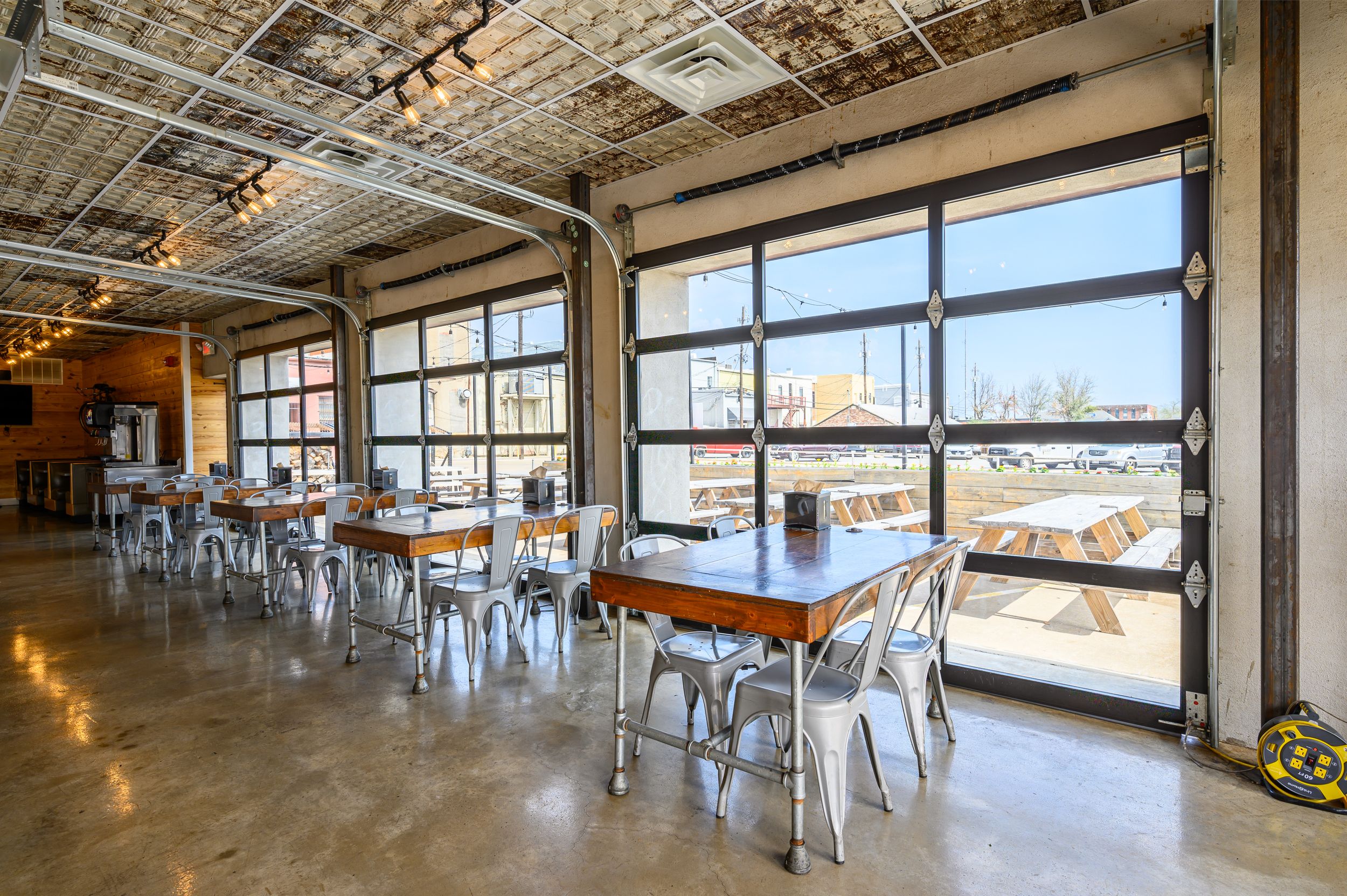 Industrial style restaurant interior with large windows, exposed ceiling, and metal chairs at wooden tables.