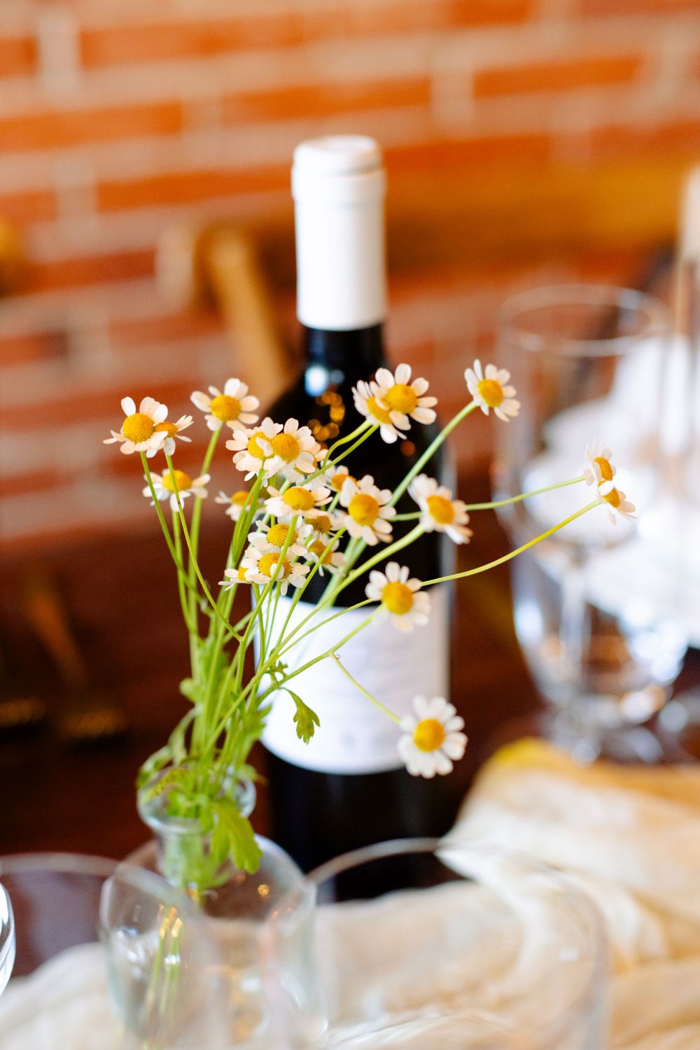 Fresh daisies in a small glass vase next to a wine bottle on a rustic wooden table.