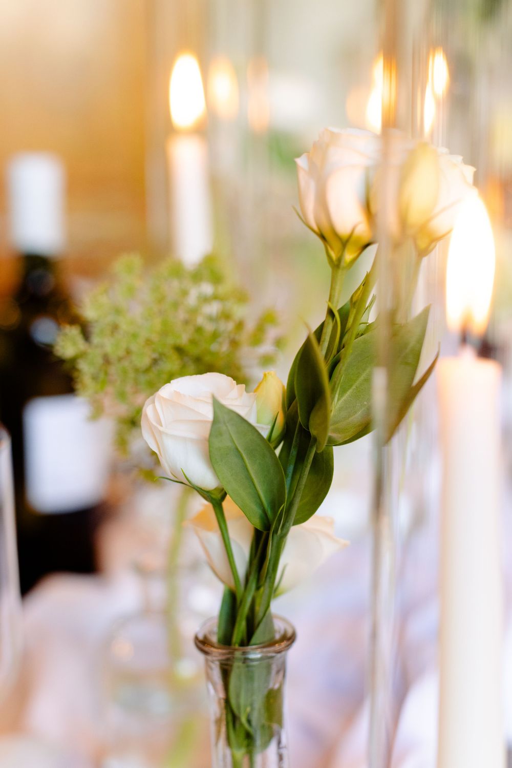 White roses and greenery in a bud vase with glowing candles creating romantic ambiance.