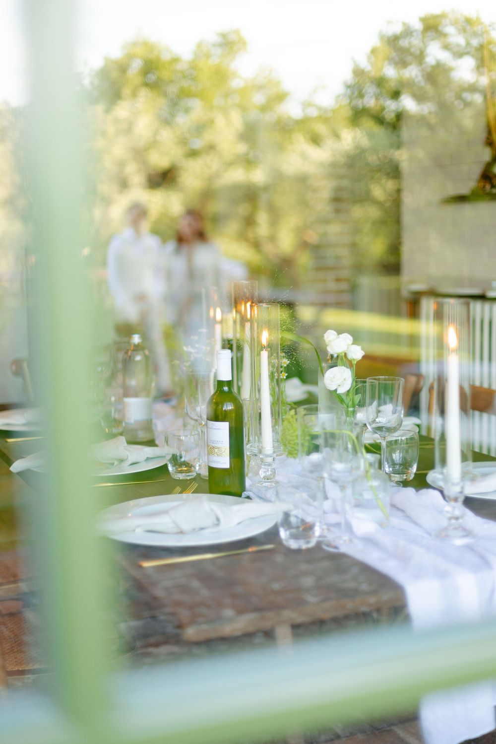 Elegant outdoor table setting viewed through window with wine glasses and white floral arrangements.