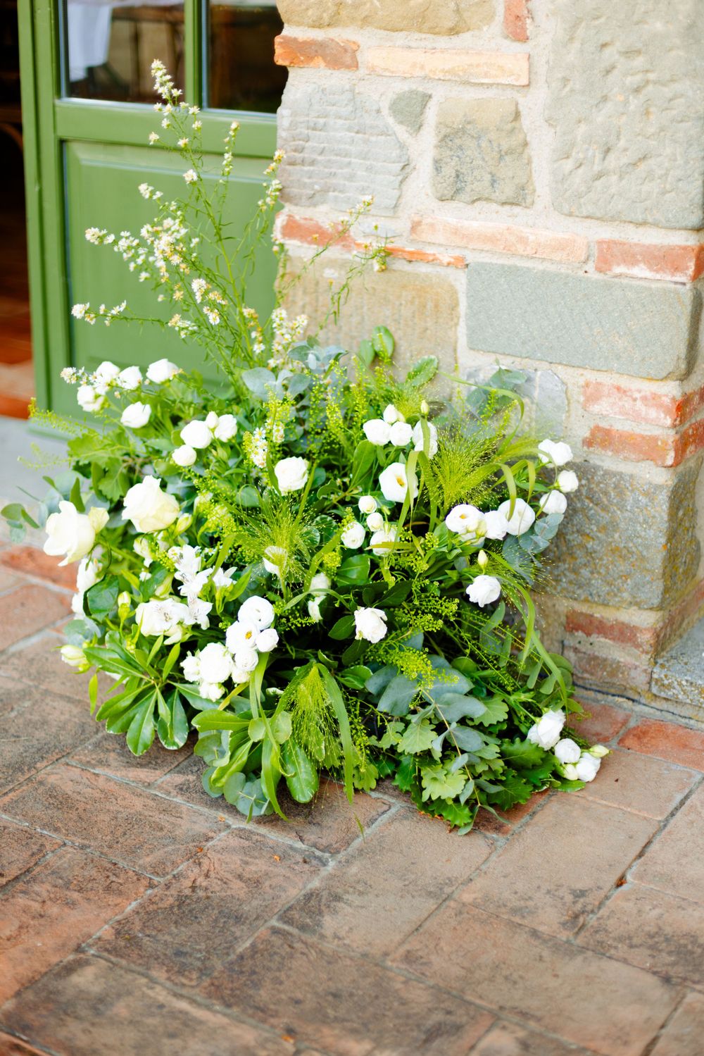 White garden flowers growing beside green door and brick wall creating charming entrance.
