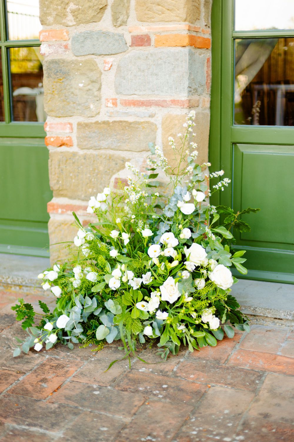 White blooming flowers and greenery arranged by green doorframe against stone wall.
