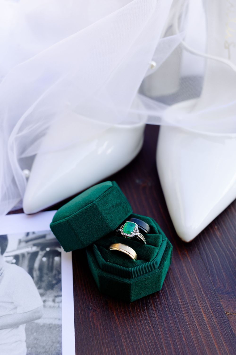 White bridal shoes and emerald green velvet ring boxes displayed on dark wood.
