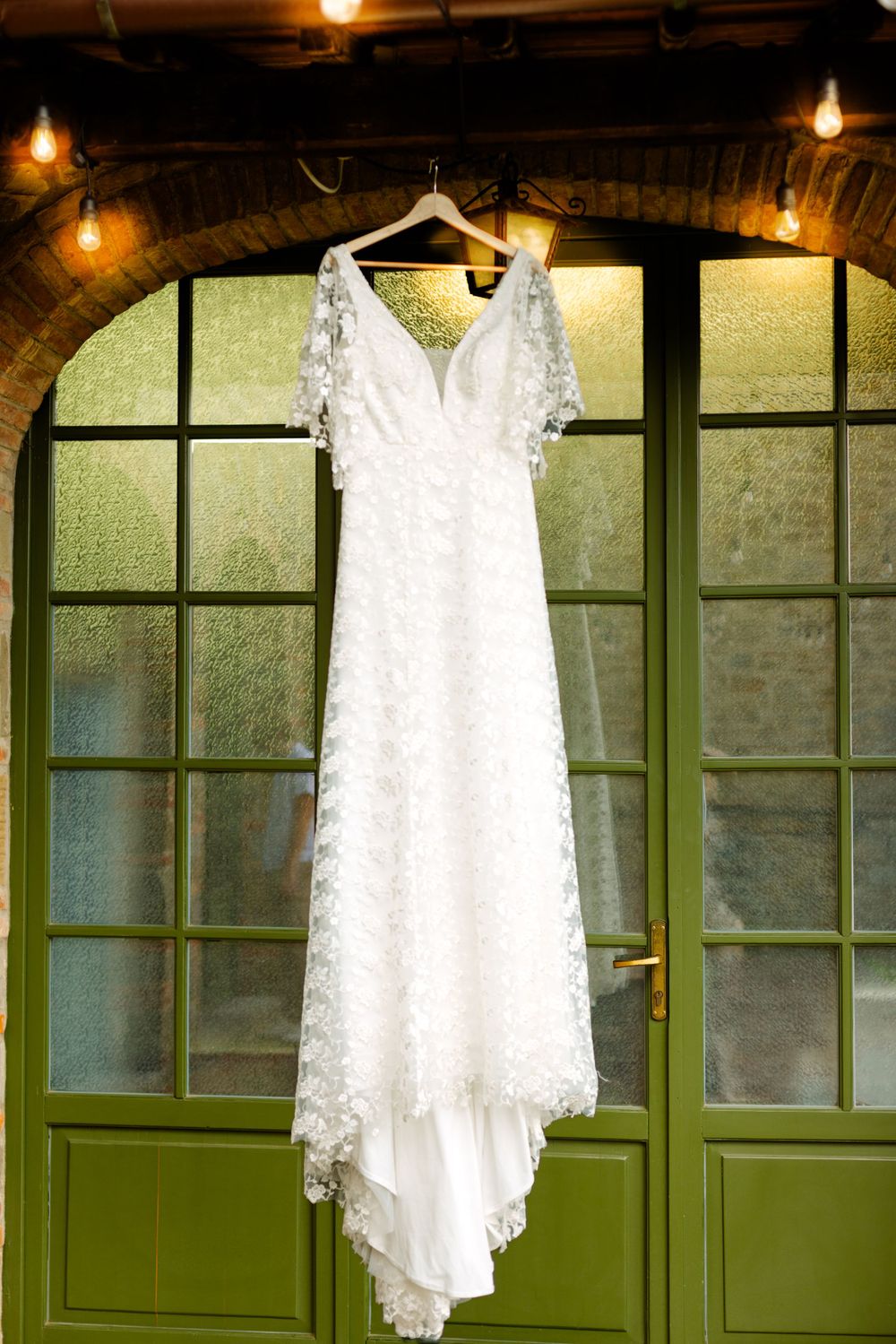 White lace wedding dress hanging against green doors with vintage lighting.