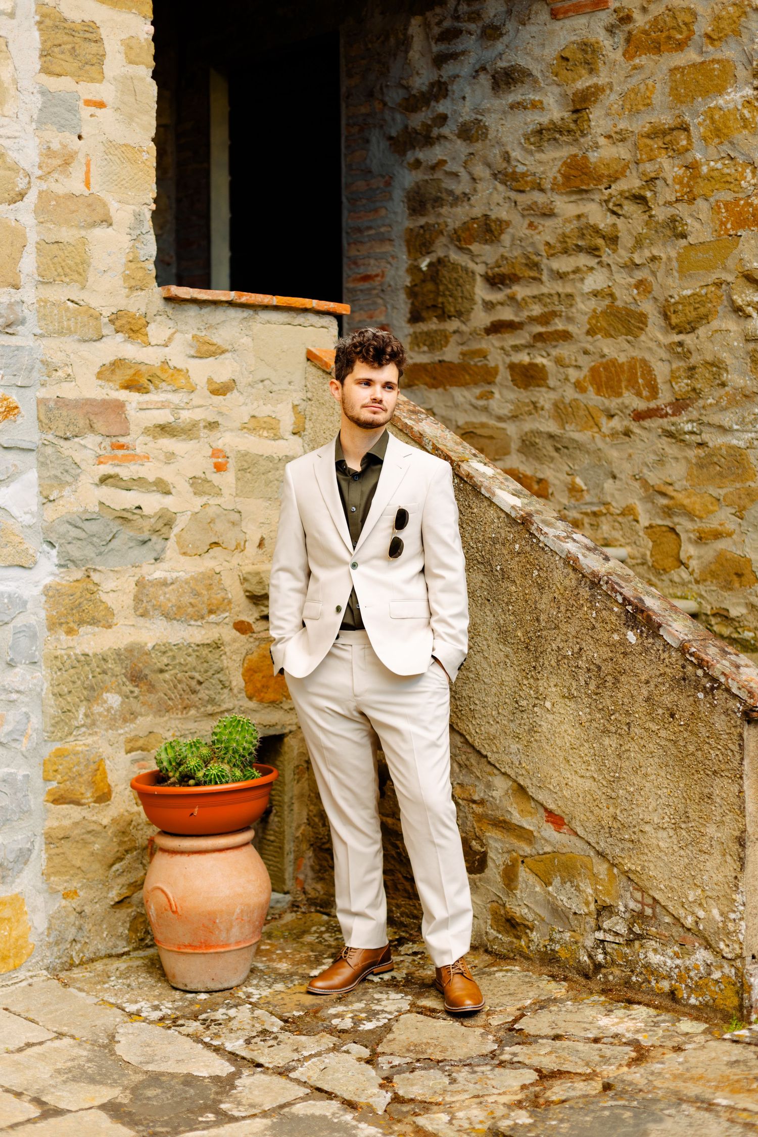 Fashionable person in light suit poses against rustic stone wall with terracotta planter.