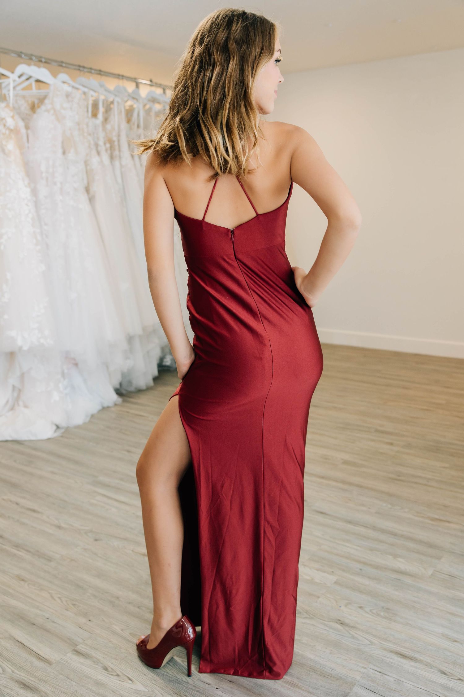 Woman in elegant burgundy satin backless gown with slit and red heels in bridal shop.