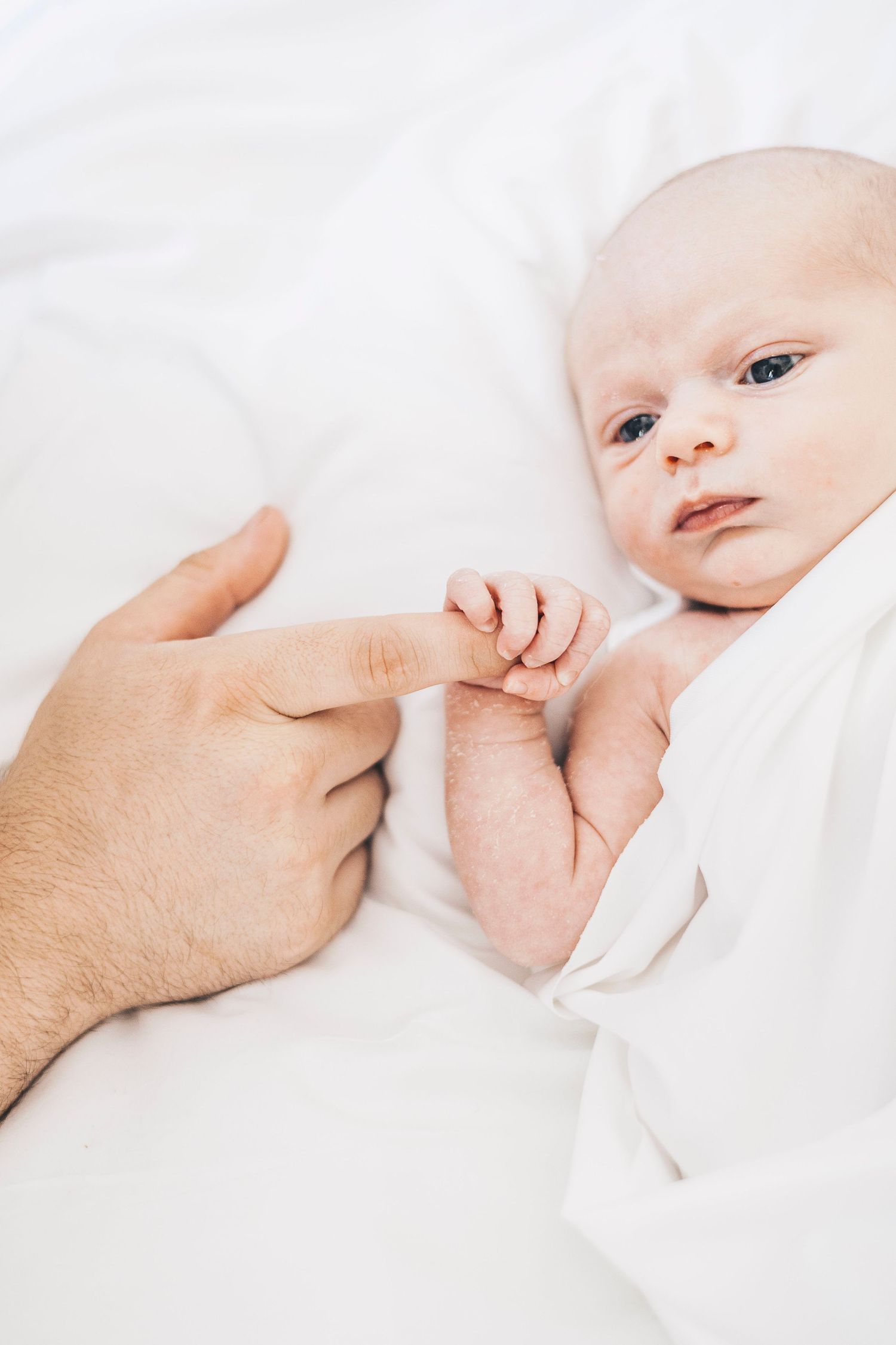 Newborn baby lying on white bedding grasps adult finger with tiny hand in tender close-up portrait.