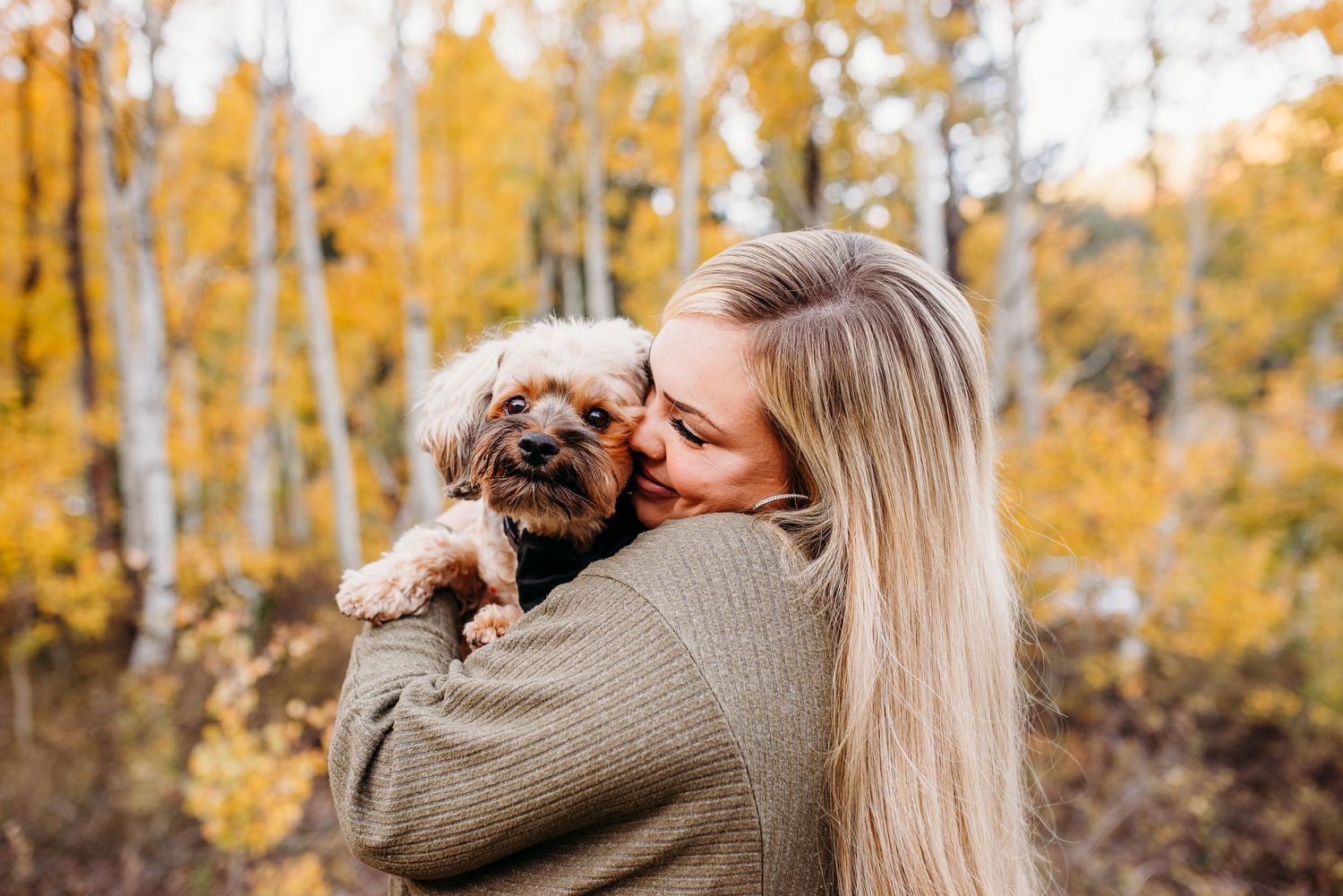 Woman in sweater cuddling small brown dog against backdrop of golden autumn trees.