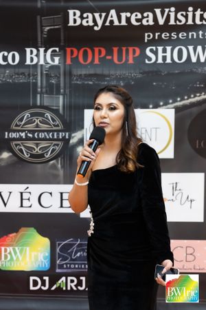 A presenter speaks into a microphone at the San Francisco BIG POP-UP SHOW with sponsor logos on the backdrop.