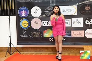 A presenter in a bright pink dress speaks on stage at a Bay Area event with sponsor logos in background.