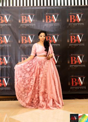 A pink floral lehenga ensemble is displayed at a formal event against a branded backdrop.