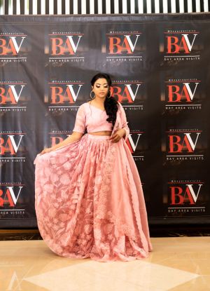 A pink two-piece lehenga outfit is showcased against a dark backdrop with BV logos.