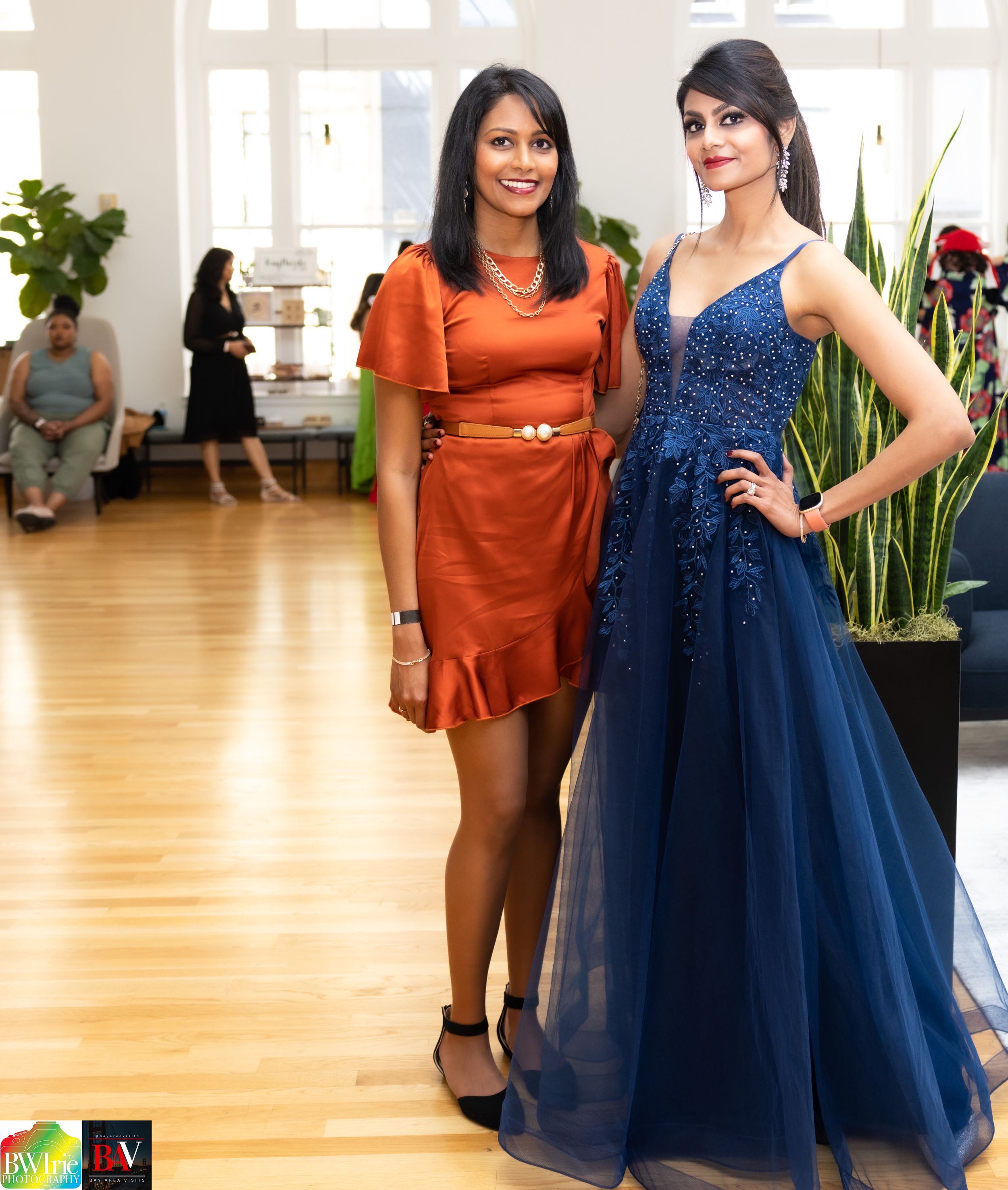 Two women pose together at an event wearing elegant dresses in orange and sparkling blue.