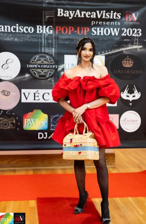 Model in dramatic red off-shoulder dress poses with straw handbag at Bay Area pop-up fashion show.